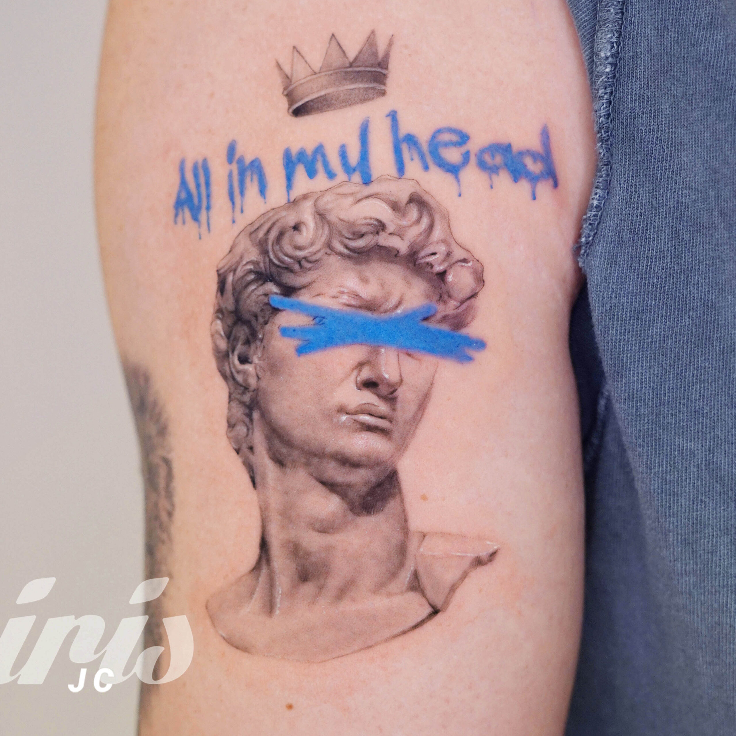 Realistic arm tattoo of a classical sculpture bust with a crown, graffiti-style blue text reading “All in my head,” and blue paint strokes covering the eyes.
Tatuaje hiperrealista en el brazo de un busto clásico con corona, texto azul que dice “All in my head” y una franja azul que cubre los ojos de la figura.