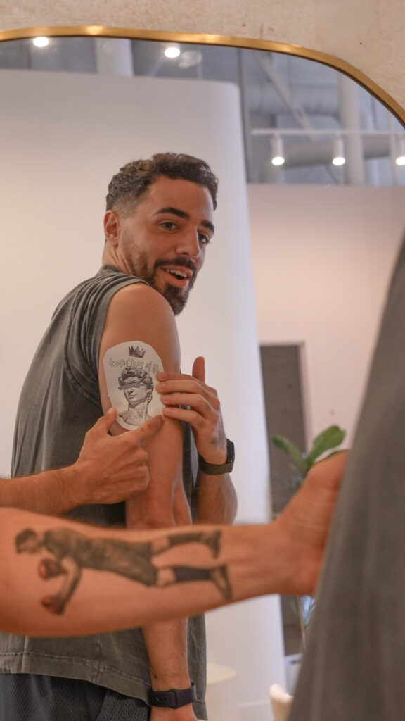 Hombre señalando un diseño de tatuaje temporal en su brazo frente a un espejo, mostrando entusiasmo y emoción.
Man pointing at a temporary tattoo design on his upper arm while looking in the mirror, showing excitement and curiosity.